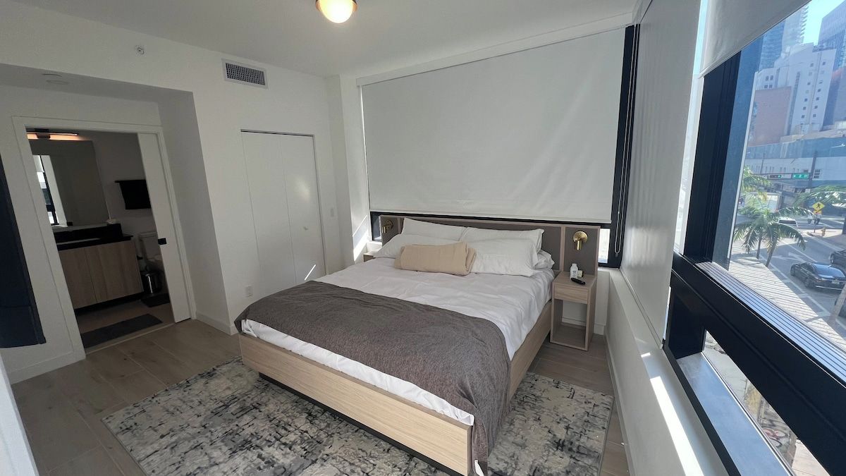 A hotel room with a bed, bedside tables, a window with a view of the city, and a door leading to another room.
