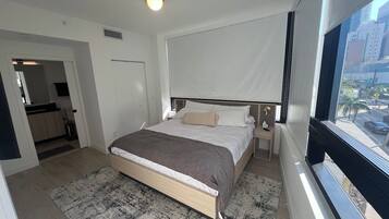 A hotel room with a bed, bedside tables, a window with a view of the city, and a door leading to another room.
