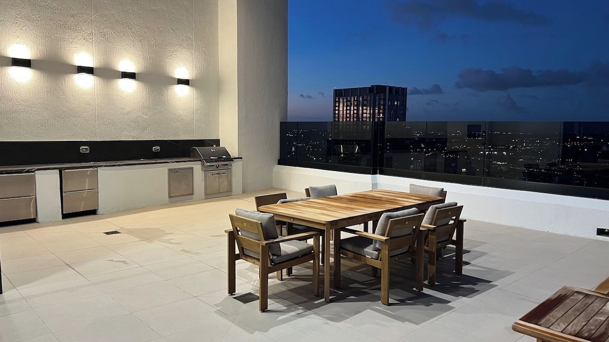 A rooftop terrace with a dining table and chairs, a built-in grill, and a city view at dusk.
