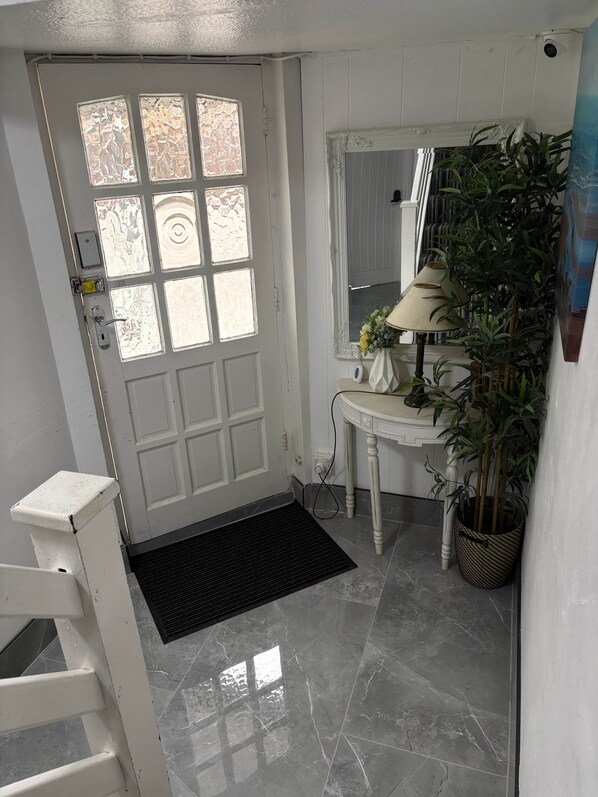 A white door with frosted glass panels, a white wall with a decorative molding, a small white table with a lamp and flowers, and a potted plant. - EasyStay Studio-Modern 2 Bedroom Family Studio Near M1, Airport & Train Station (Luton)