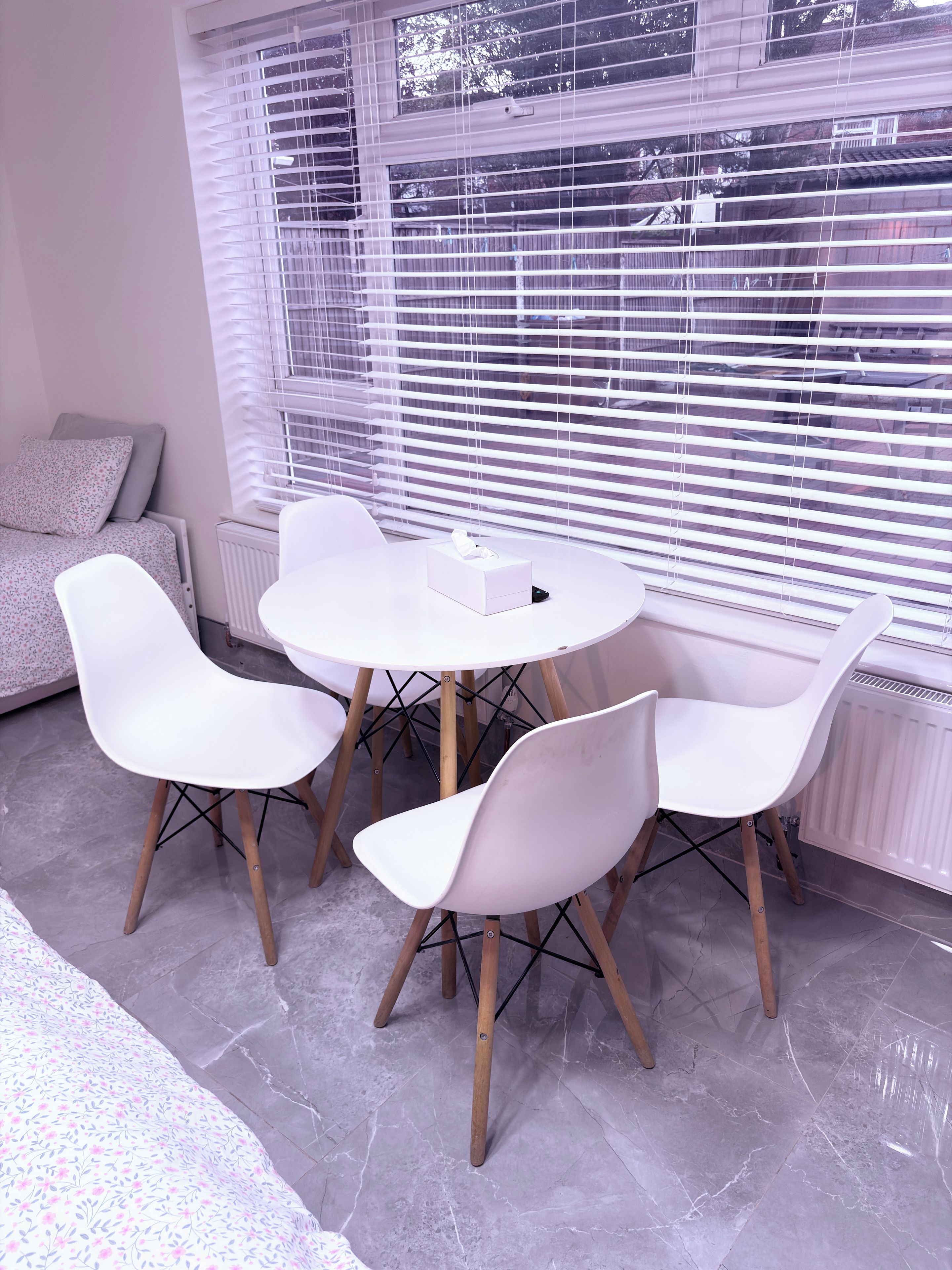 A modern room with a round table, four white chairs, a radiator, and a window with blinds.