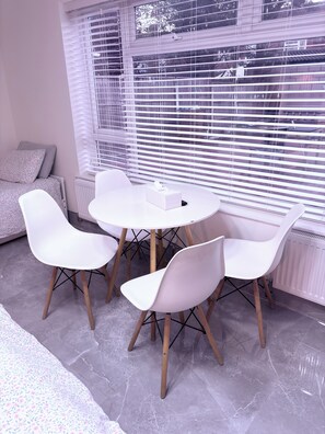 A modern room with a round table, four white chairs, a radiator, and a window with blinds. - EasyStay Studio-Modern 2 Bedroom Family Studio Near M1, Airport & Train Station (Luton)