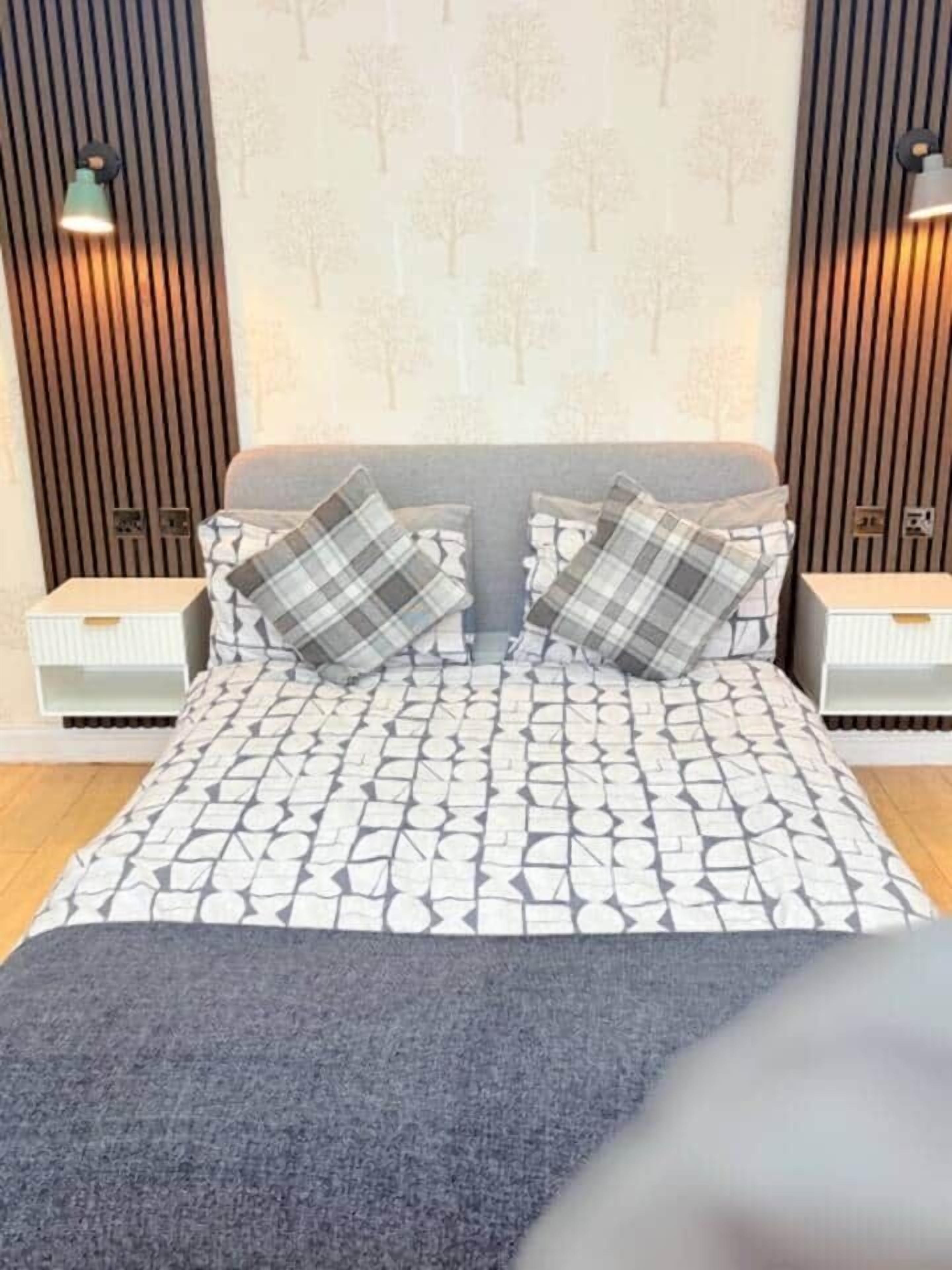 Une chambre moderne avec une couvre-lit à motifs, deux tables de chevet et un mur orné de lattes de bois verticales et d’un motif représentant un arbre.