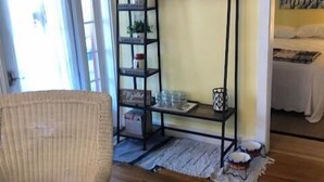 A room with a wicker chair, a wooden floor, a metal shelving unit with various items, and a bed with white bedding. - 317 Maple Unit 2 in Lakeside (Lakeside)