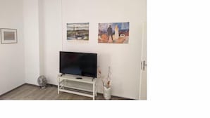 A room with a flat-screen TV on a white stand, a small fan, and two paintings on the wall.