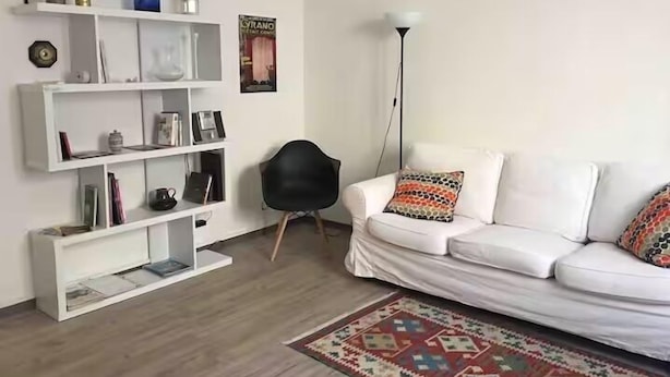 A living room with a white sofa, a black chair, a patterned rug, and a white shelving unit with various items.