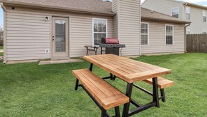 A backyard with a wooden picnic table and benches, a barbecue grill, and a house with beige siding. - Quiet Stay w Workspace, Fast Wi-Fi & Parking (Fishers)