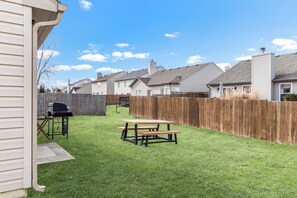 Une cour arrière avec une table de pique-nique et un barbecue, entourée d’une clôture en bois et avec des maisons en arrière-plan.