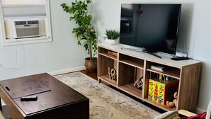 A living room with a flat-screen TV on a wooden stand, a dark coffee table, a wooden chair, and a small bookshelf with children's toys.