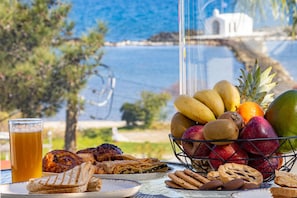 A table with a fruit basket, pastries, and a glass of beer, overlooking a scenic coastal view. - ARETI LUXURY APARTMENT (Apokoronas)