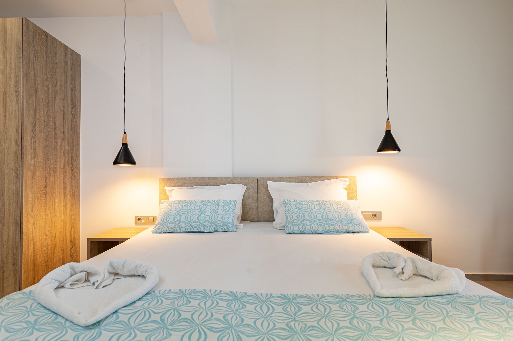 A neatly made bed with patterned pillows and a matching blanket, two bedside lamps, and a wooden wardrobe.
