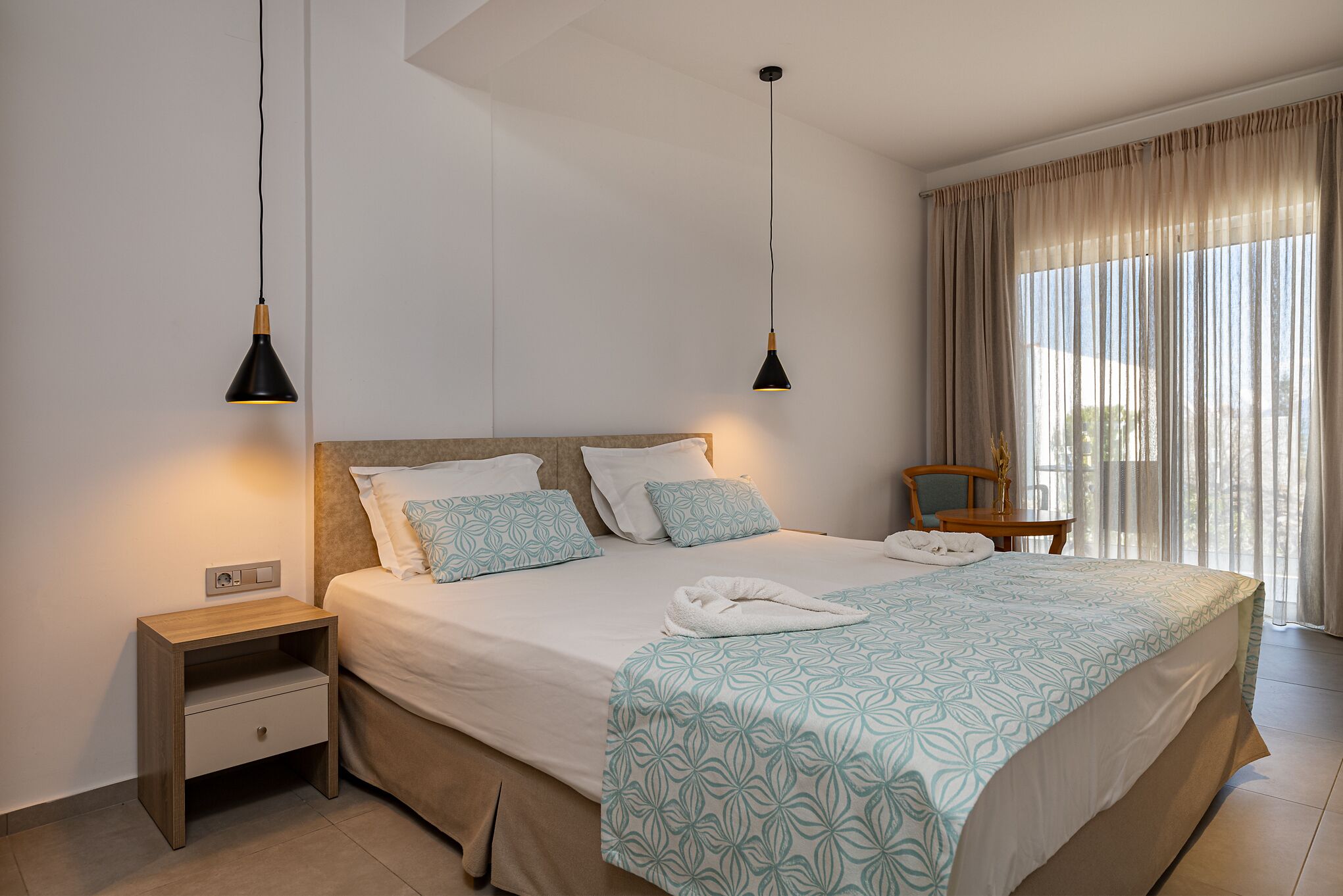 A neatly made bed with a patterned bedspread, a bedside table with a lamp, and a chair near a window with curtains.