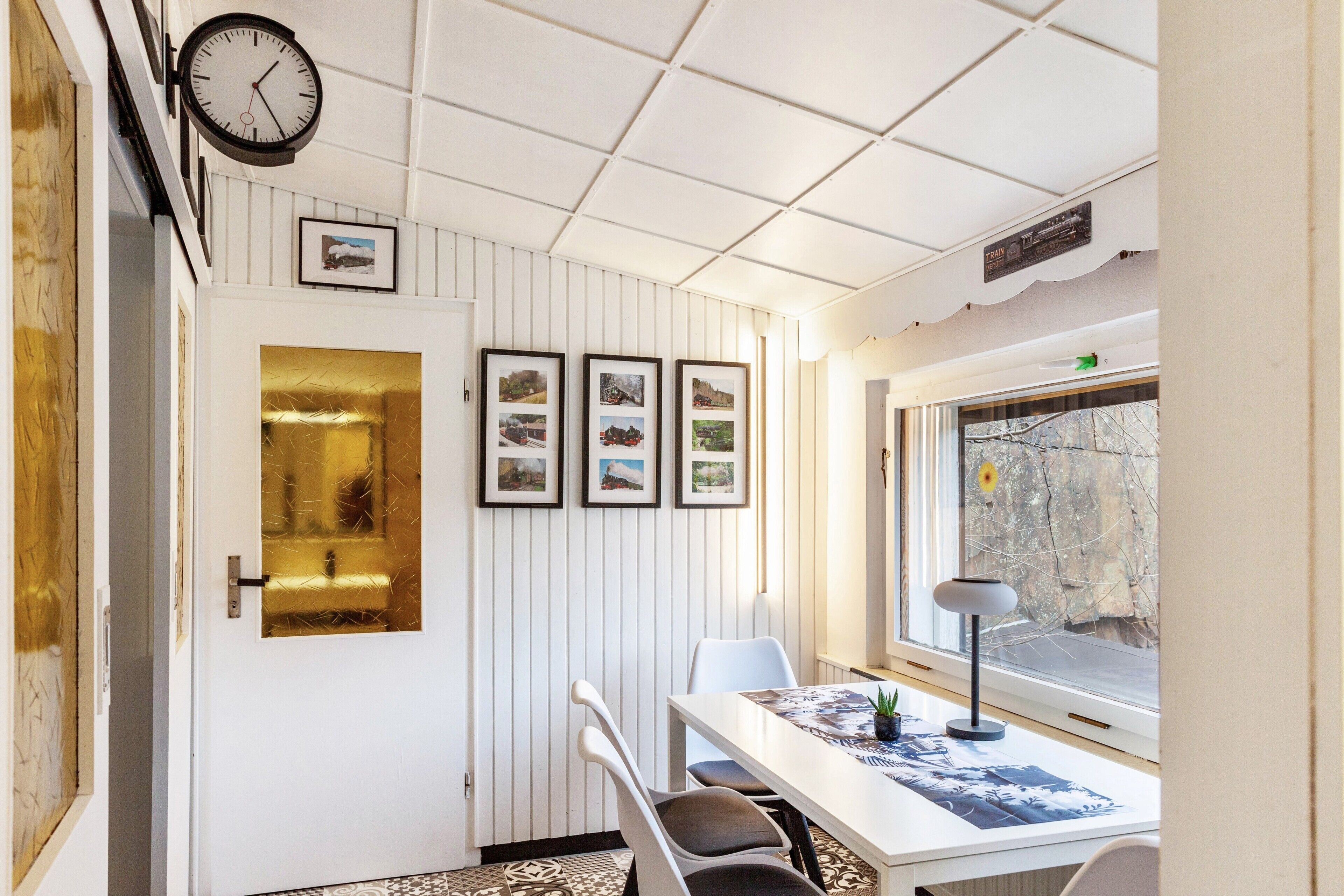 A room with a white wall, a clock, framed pictures, a table with chairs, and a window with a view.