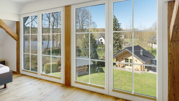 A room with large windows, wooden flooring, and a view of a house and trees. - Vogtlandstadl – Modern Lakeside Holiday Home in Vogtland, Spacious & Comfortable (Bad Brambach)