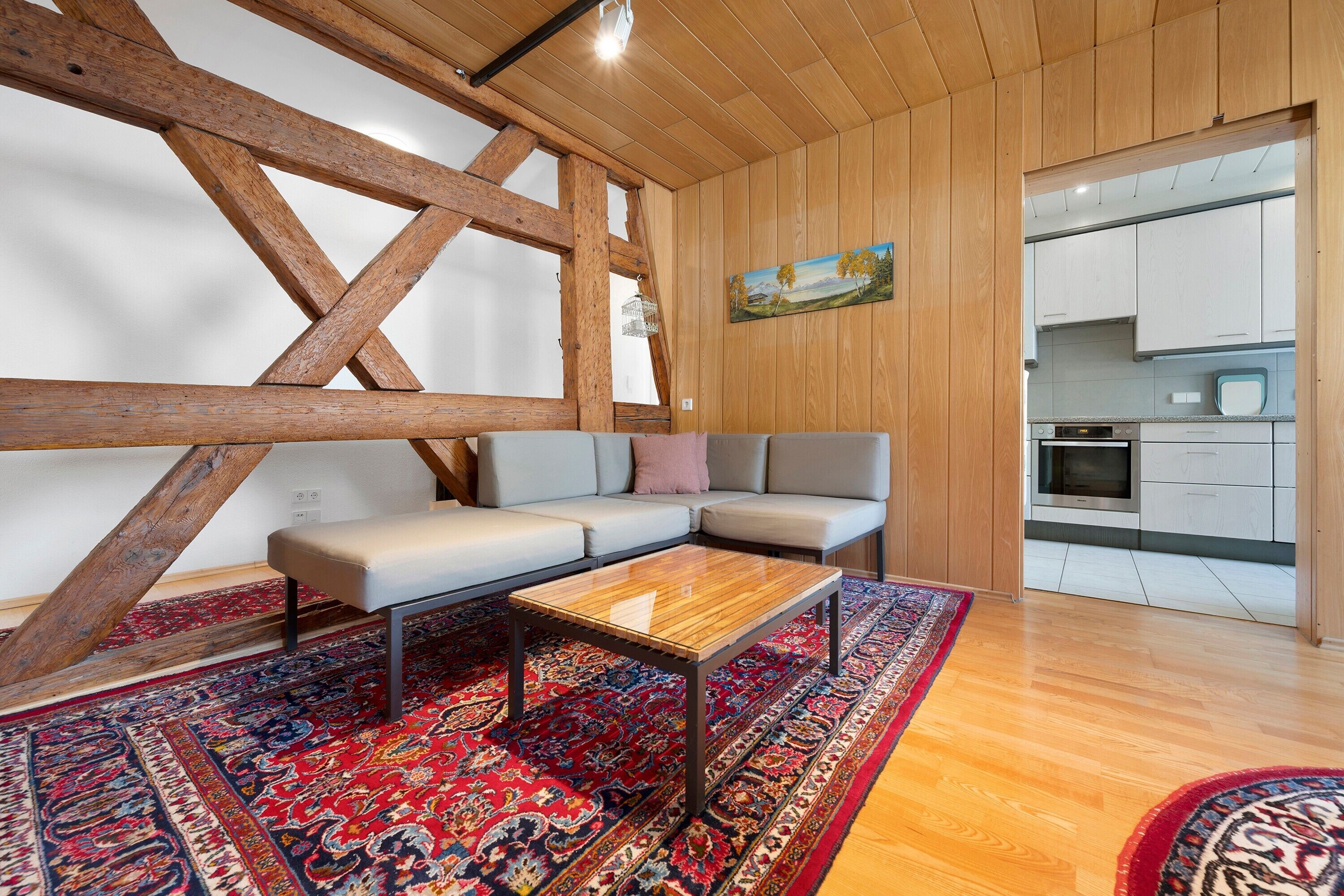 A modern living room with a wooden ceiling, a sofa, a coffee table, and a kitchen visible through an open door.