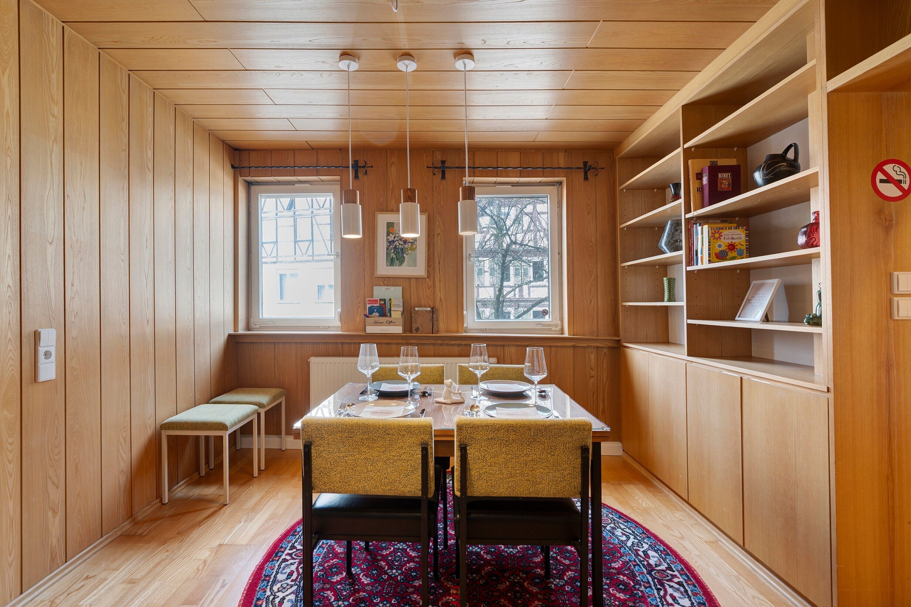 Un coin repas avec une table en bois, des chaises et un tapis. On y trouve des étagères remplies de livres et un panneau indiquant qu'il est interdit de fumer.