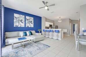 A modern living room with a blue accent wall, a white sofa, a coffee table, and a dining area with bar stools. - Disney Min Away - Pool, Patio & Playground (Kissimmee)