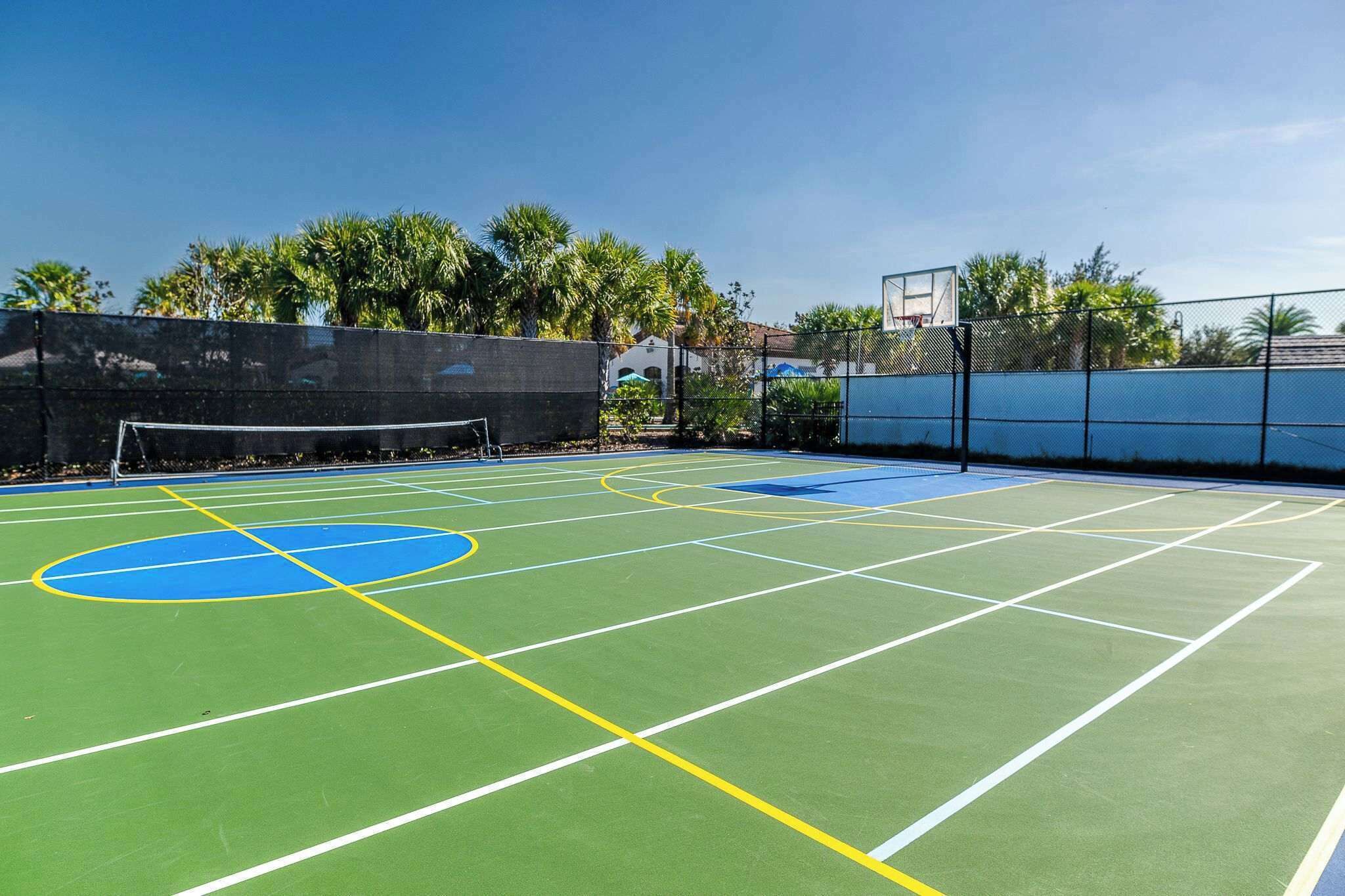An outdoor tennis court with a net, surrounded by a fence and palm trees.