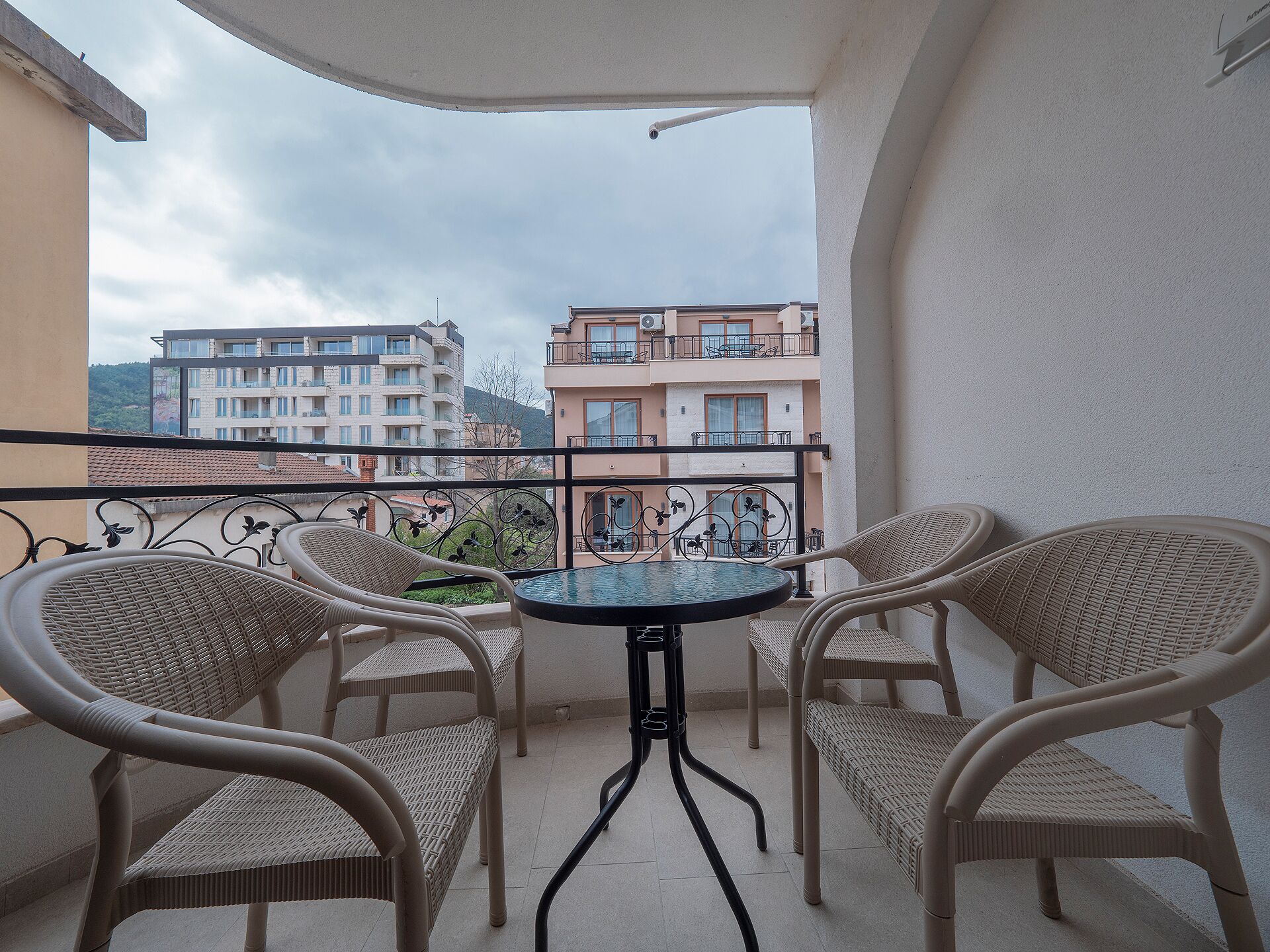 A balcony with wicker chairs and a small table, overlooking a cityscape with buildings and a mountain in the background.