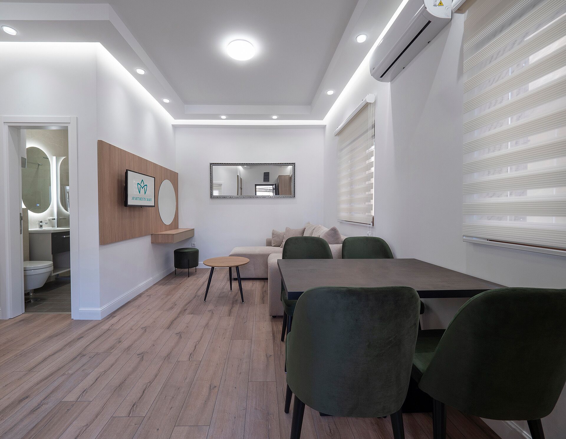 Una sala de reuniones moderna con suelo de madera, una mesa redonda y sillas verdes. Hay un sofá y un espejo grande en la pared.