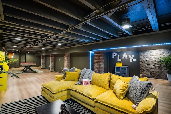 Un área lounge moderna con mesa de billar, un sofá amarillo y una pared con la palabra 'PLAY'.