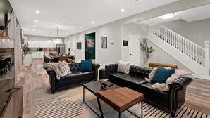 Un vaste salon comprenant un ensemble de canapés en cuir noir, une table basse en bois et un ventilateur de plafond. On aperçoit un escalier au fond de la pièce, menant à un niveau supérieur.