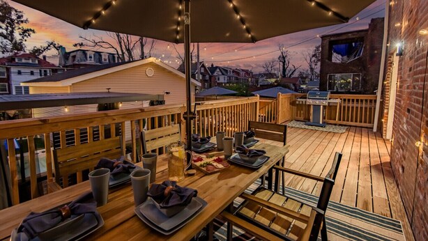 Une terrasse extérieure avec une table en bois dressée pour les repas, entourée d’une balustrade en bois, et des guirlandes lumineuses suspendues au-dessus.