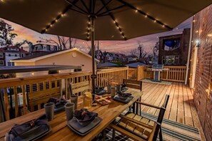An outdoor deck with a wooden table set for dining, surrounded by a wooden railing, and string lights overhead. - Family Luxury Getaway: Hot Tub, Sauna & Game Room (St. Louis)