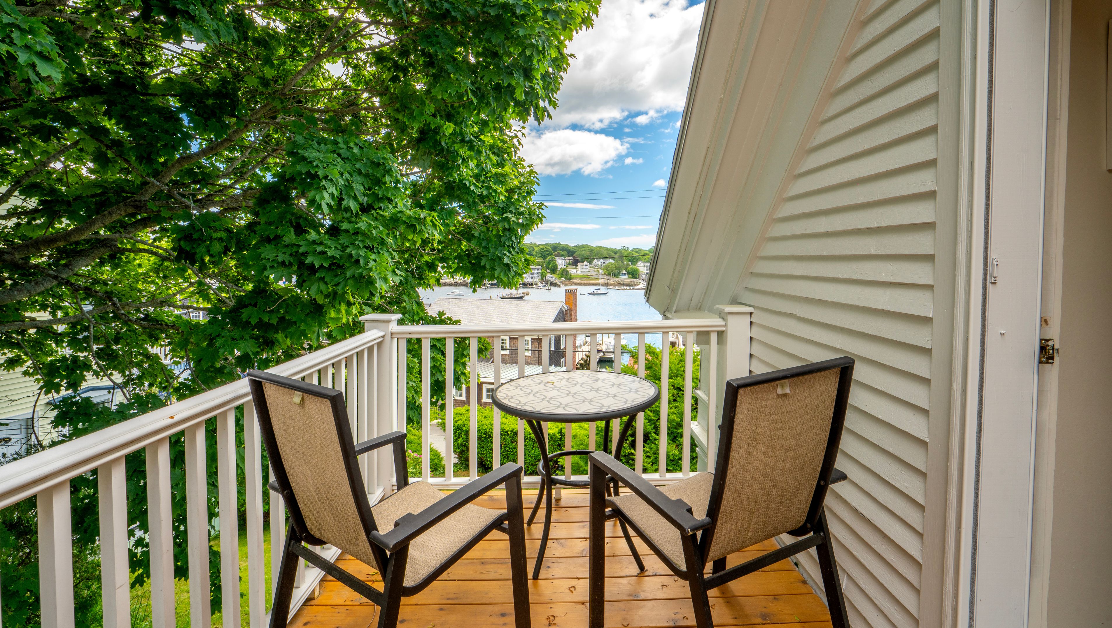 A wooden deck with a table and two chairs, overlooking a body of water with boats and buildings in the distance.