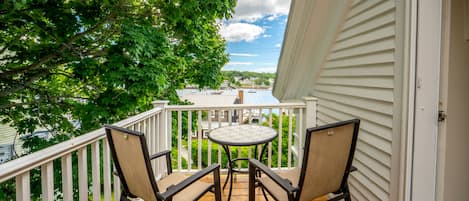 A wooden deck with a table and two chairs, overlooking a body of water with boats and buildings in the distance.