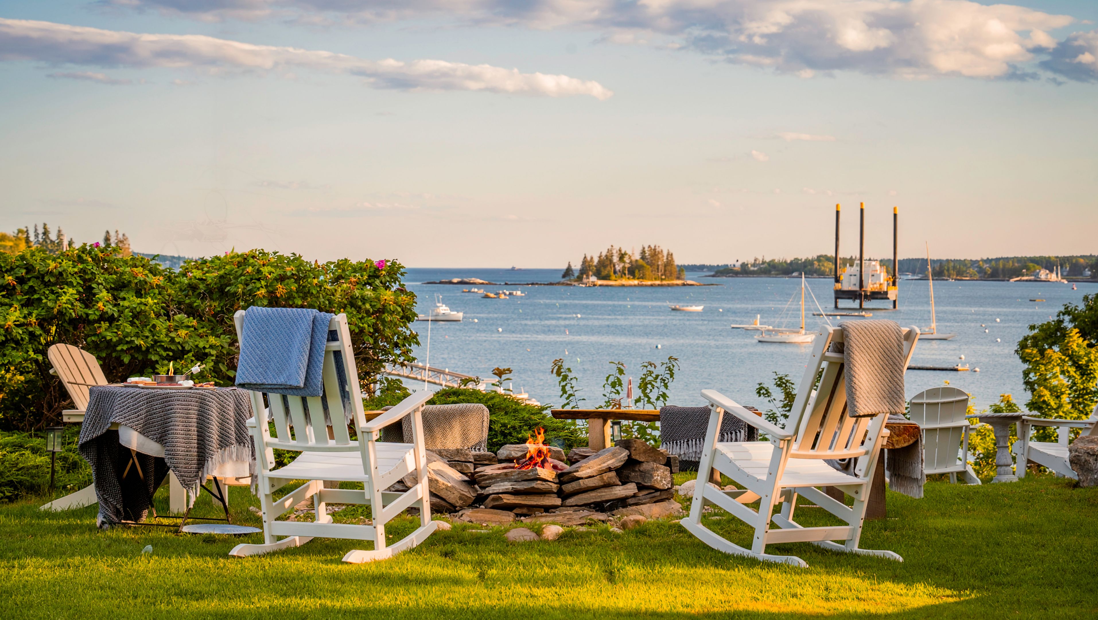 A serene lakeside setting with two rocking chairs facing a calm body of water, a fire pit in the center, and a clear view of boats and distant land.