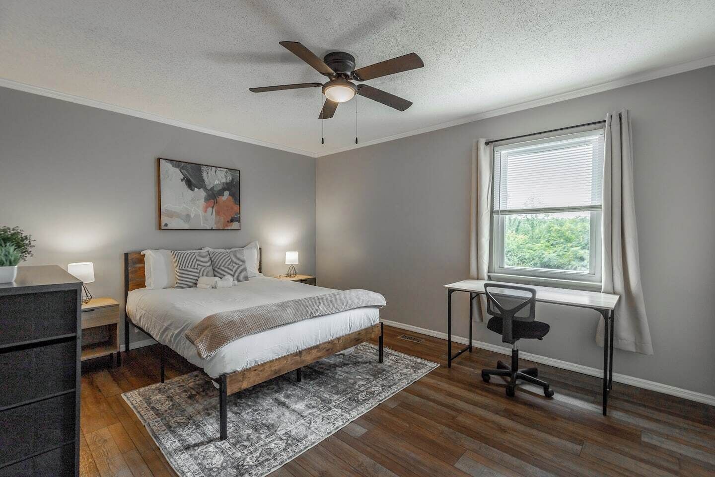 Une chambre à coucher comprenant un lit, des tables de chevet, un bureau, un ventilateur de plafond, une fenêtre avec des rideaux et un tableau accroché au mur.