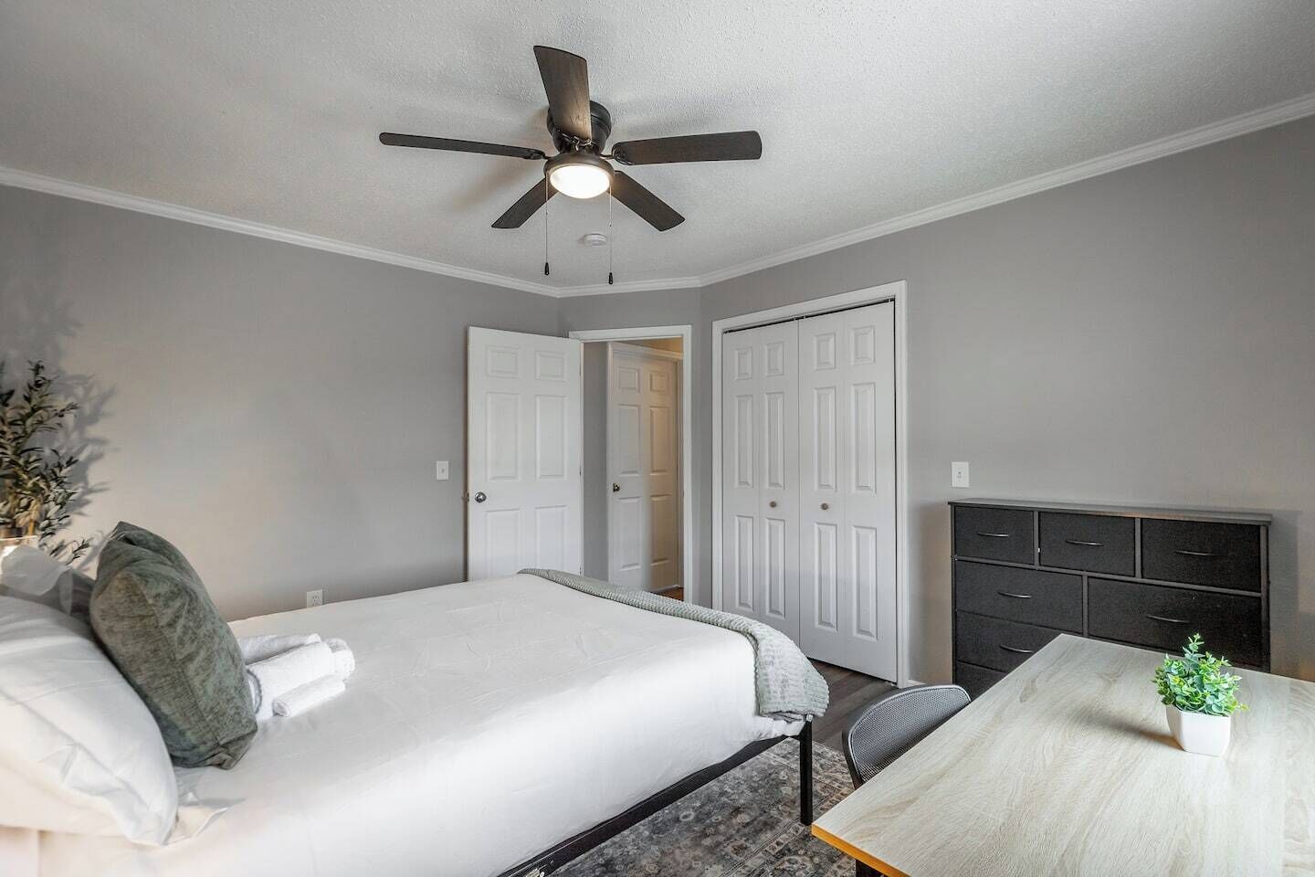A bedroom with a bed, a desk, a chair, a ceiling fan, and a dresser.