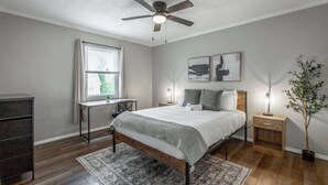 A bedroom with a bed, a desk, a chair, a ceiling fan, a window with curtains, and a plant.