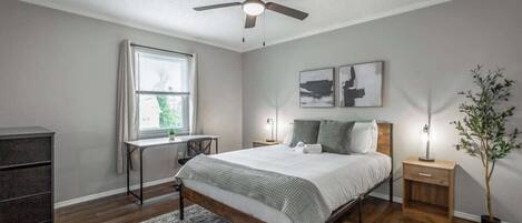 Une chambre à coucher comprenant un lit, un bureau, une chaise, un ventilateur de plafond, une fenêtre avec des rideaux et une plante.