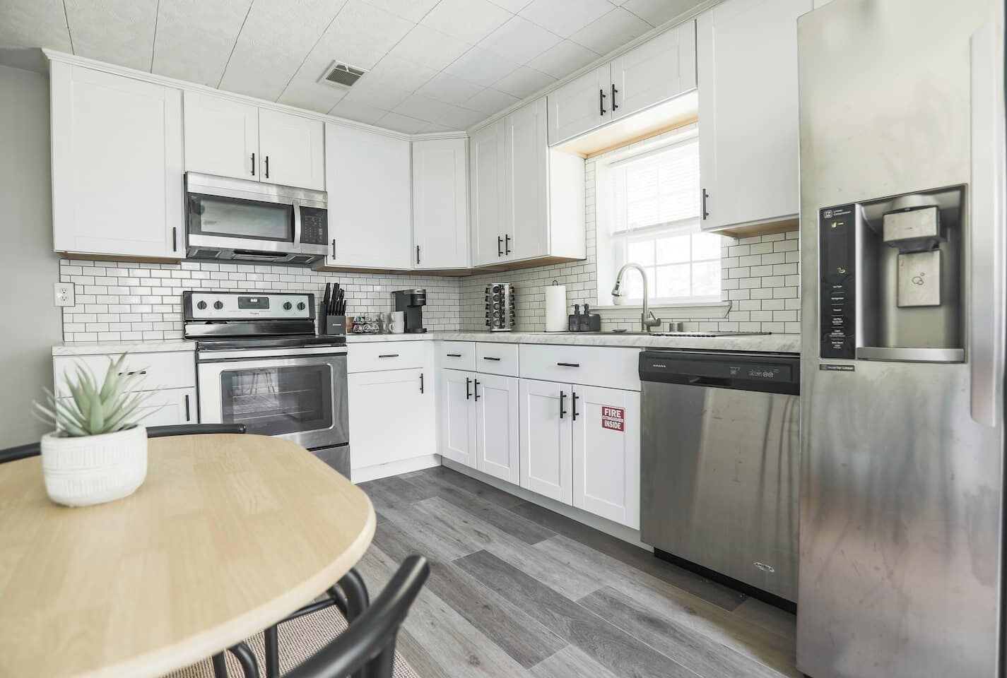 A modern kitchen with white cabinets, stainless steel appliances, a wooden dining table, and a potted plant.
