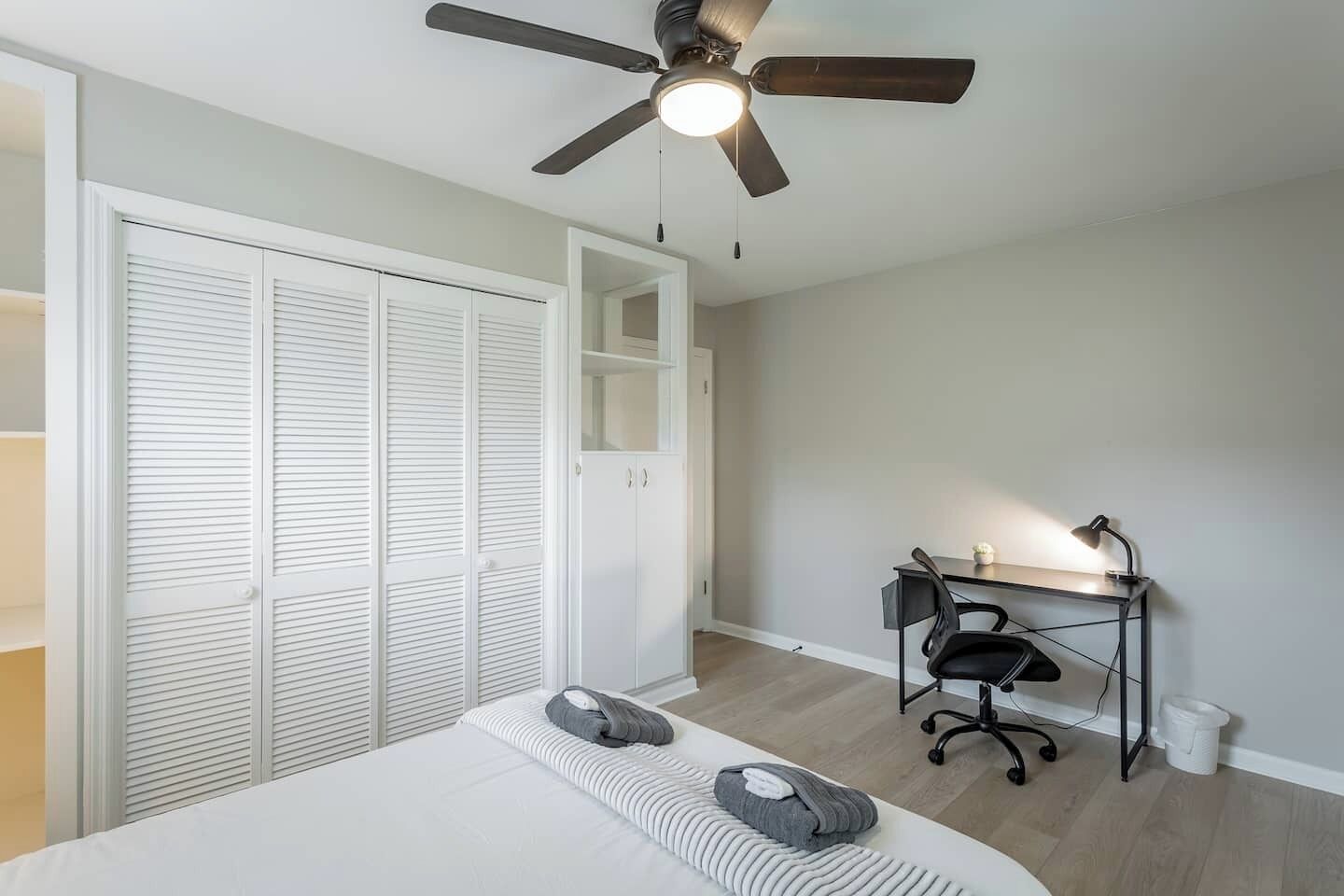 Une chambre à coucher comprenant un lit, un bureau avec une chaise, un ventilateur de plafond et une armoire avec des portes coulissantes.