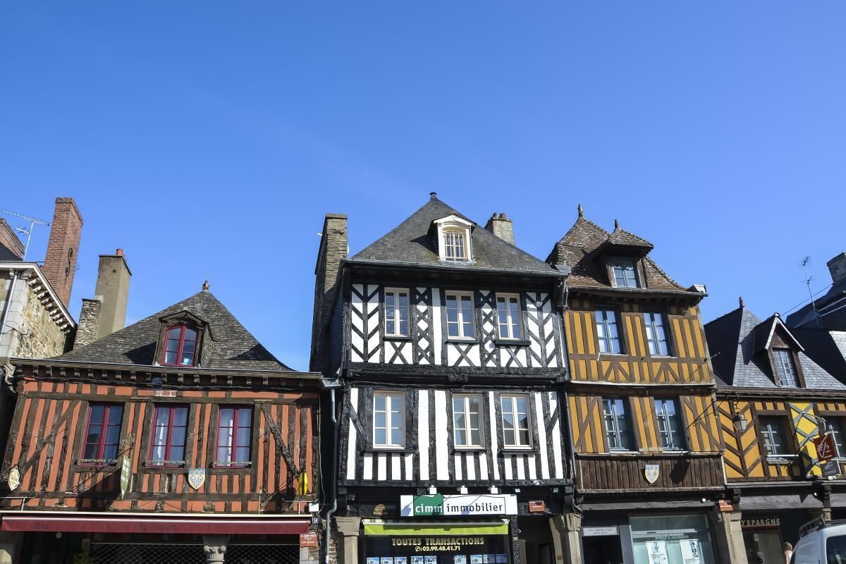 A row of traditional half-timbered buildings with a mix of black and white facades, a visible storefront with a sign for 'cim' and 'immobilier', and a clear blue sky above.