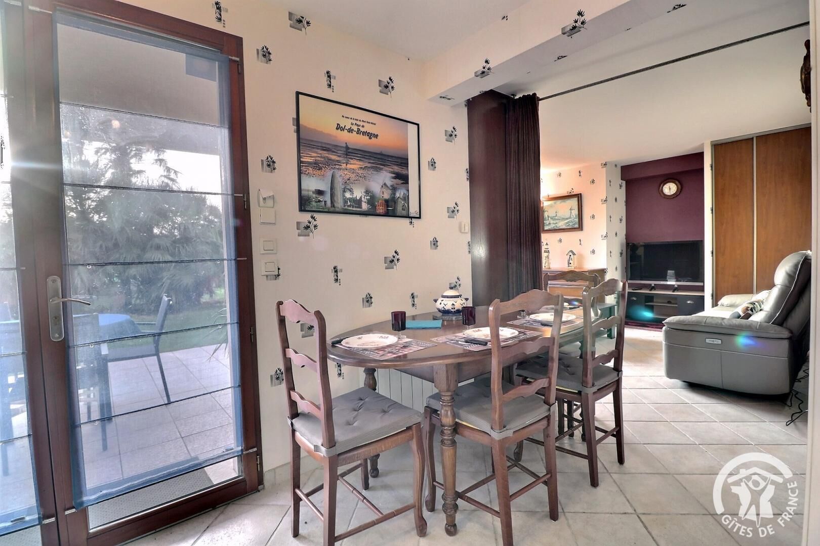 Un coin repas avec une table et des chaises en bois, une porte vitrée donnant sur l’extérieur, et un espace salon avec un canapé et un téléviseur.