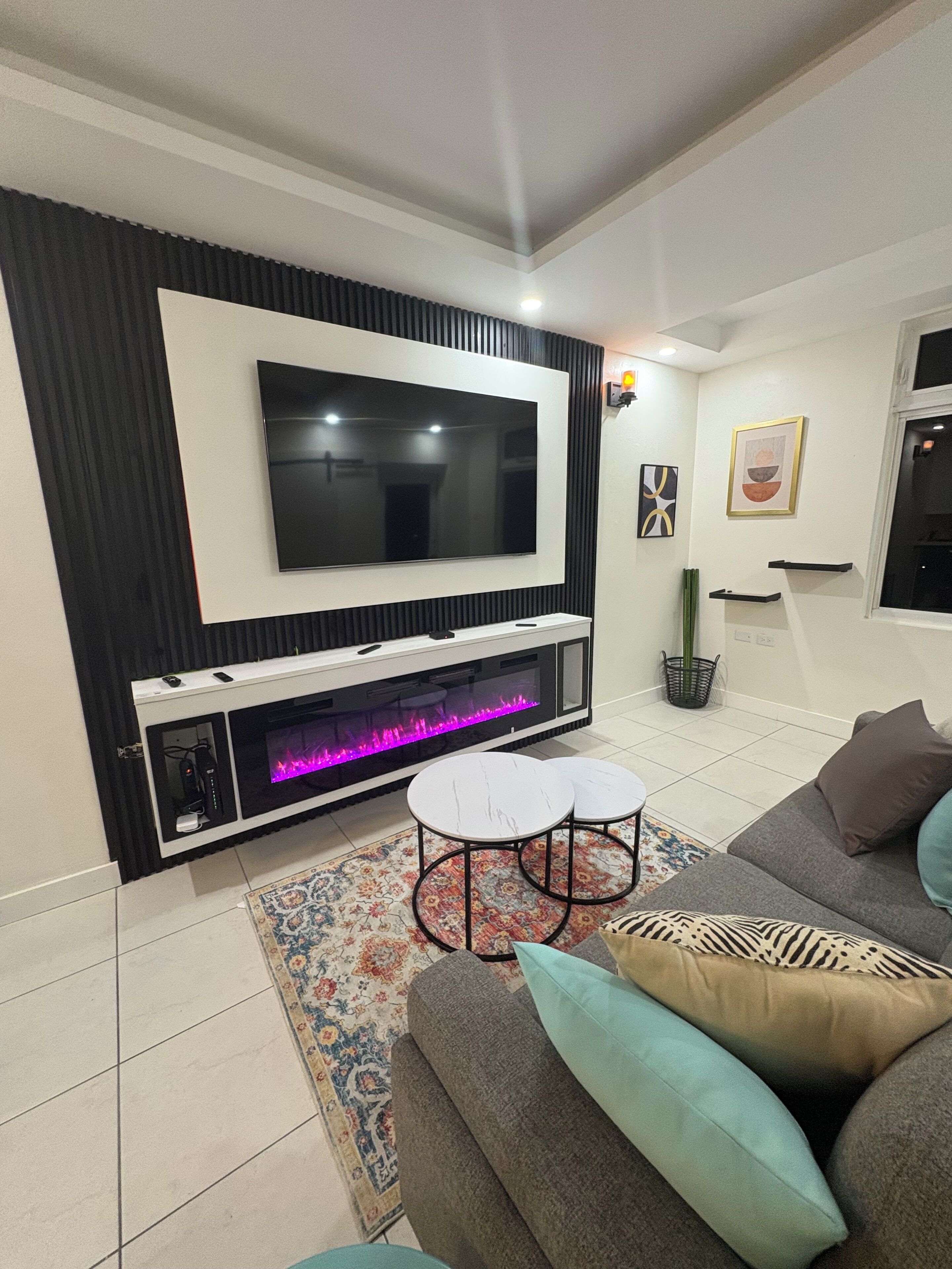 Een moderne woonkamer met een flatscreen televisie aan een zwart-wit muur, een open haard met glazen deur, een grijze bank met kussens en een kamerkleed met dessin.