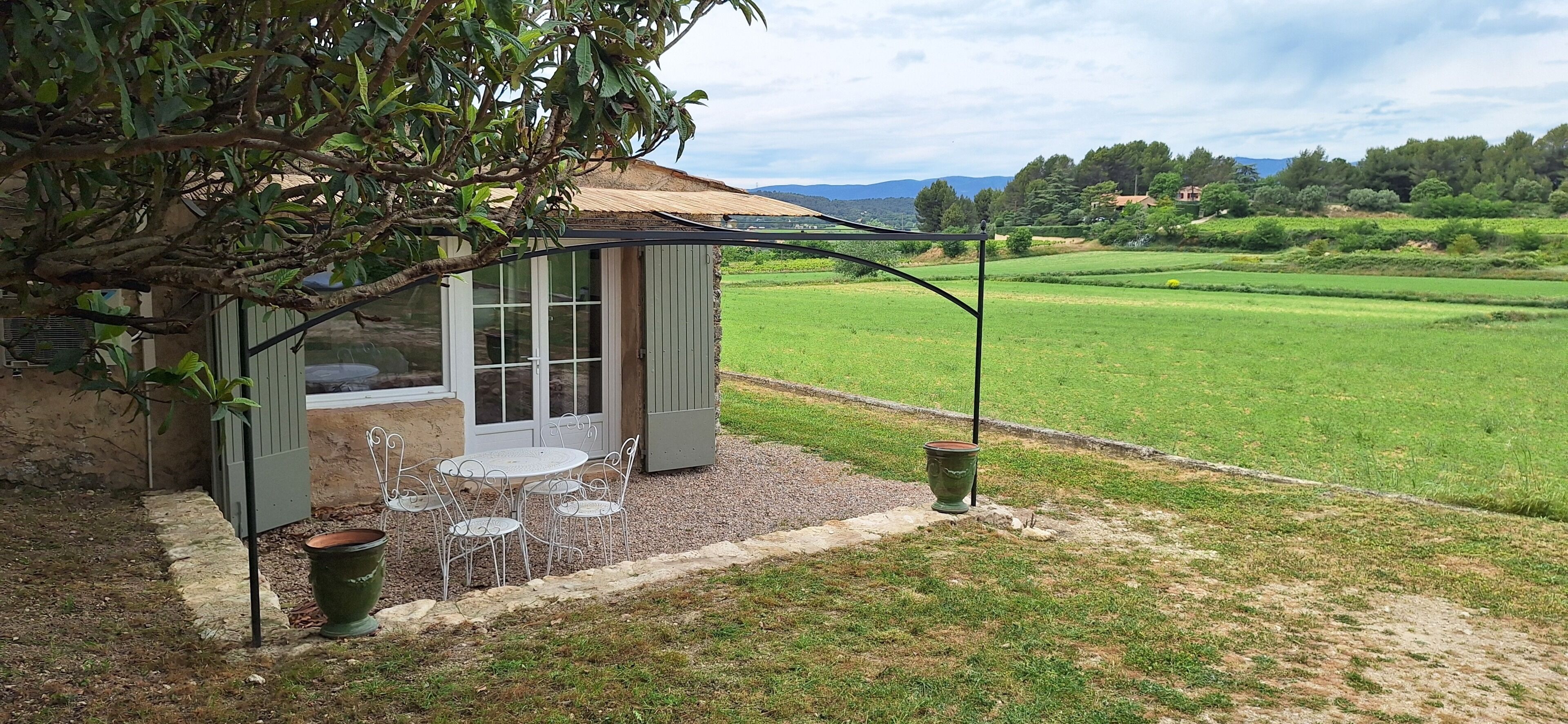 A small, open-air structure with a glass door, a table and chairs, and a view of a green field.