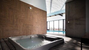 Un spa moderno con suelo de madera, una bañera grande y vistas a una piscina a través de amplios ventanales.