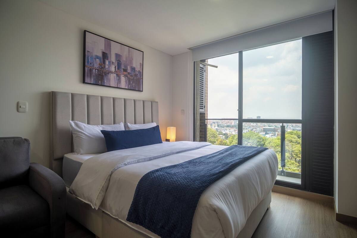 Une chambre d’hôtel moderne avec un grand lit, une tête de lit, une vue sur la ville à travers une grande fenêtre et un fauteuil.