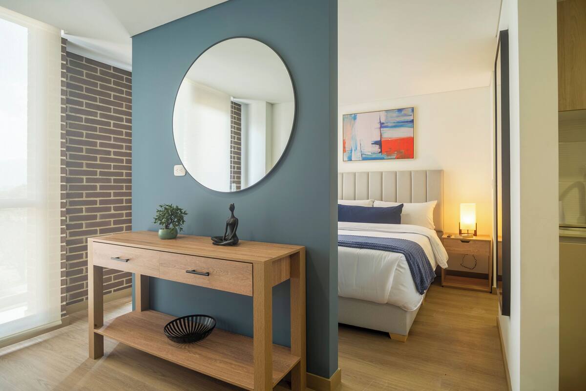Une chambre moderne avec une table de chevet en bois, un miroir rond, un lit avec une literie blanche et une table de nuit avec une lampe.