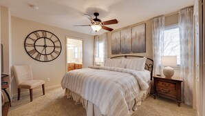Un dormitorio con un reloj de pared grande, una cama con edredón blanco, una mesita de noche con lámpara y un ventilador de techo.