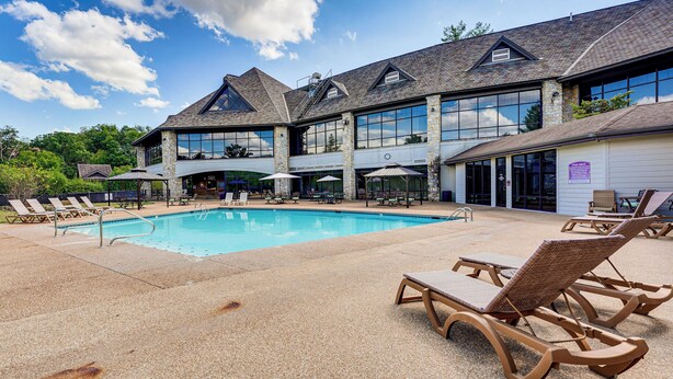 Una piscina con sillones y sombrillas, un porche cubierto y un edificio con ventanas amplias y fachada de piedra.