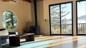 A spacious living room with a large fireplace, a round window, and a balcony with a view of trees. - Epic views, modern luxury, private retreat in West Marin on hilltop 2 acre (Lagunitas-Forest Knolls)