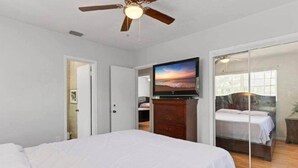 Un dormitorio con cama, televisor en una cómoda, ventilador de techo y vista a otra habitación por una puerta de vidrio.