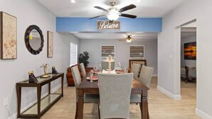 Un comedor con ventilador de techo, un letrero que dice 'Believe', obras de arte enmarcadas y una mesa de comedor preparada para cuatro.