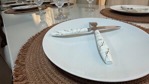 Een eettafel gedekt met witte borden, bestek en kristallen glazen, met een geweven placemat en een uitzicht op een stenen muur en gordijnen op de achtergrond.